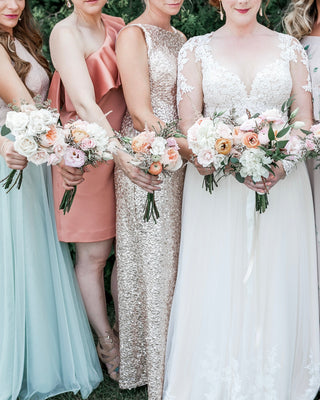 Bride with bridesmaids in various pastel dresses holding bouquets outdoors.