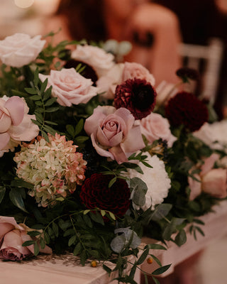 A floral centrepiece arrangement featuring a variety of flowers including pink roses, gerbera daisies, and green foliage, displayed in a wooden structure.