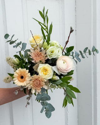 Hand-tied bouquet of flowers (candlelight roses, blush ranunculus, blush gerbera daisy, cream and green lisianthus, white tulips, ruscus, eucalyptus) in blush pink and cream colours held against a white wooden door background