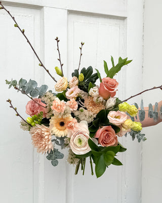 Hand-tied bouquet of flowers (latte roses, blush ranunculus, blush gerbera daisy, rose gold chrysanthemum, green, lisianthus, ruscus, eucalyptus, cherry blossom branches) in blush, latte and apricot colours held against a white wooden door background