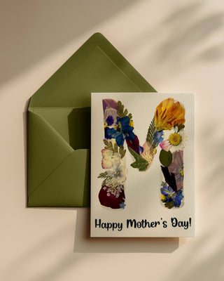 Floral card with an "M" letter made of real pressed flowers and 'Happy Mother's Day!' text with green envelope on a beige background