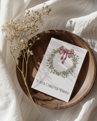 Card with a pink bow and Christmas wreath on a wooden plate with dried flowers, all on a soft white fabric background.