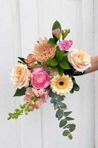 Hand-tied bouquet of flowers (apricot roses, pink ranunculus, blush gerbera daisy, soft pink snap dragon, pink freesia, ruscus, eucalyptus) in pink, blush and apricot colours held against a white wooden door background
