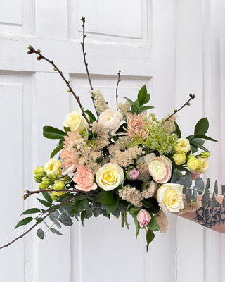 Hand-tied bouquet of flowers (candlelight roses, blush ranunculus, latte chrysanthemums, cream and green lisianthus, pink tulips, ruscus, eucalyptus) in blush pink and cream colours held against a white wooden door background