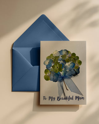 Greeting card with blue and green floral design using real pressed flowers and 'To My Beautiful Mom' text, paired with a blue envelope on a beige background.